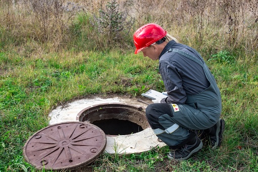 A male plumber in overalls and a helmet near a water well writes down the measurements taken and the readings of the water meter